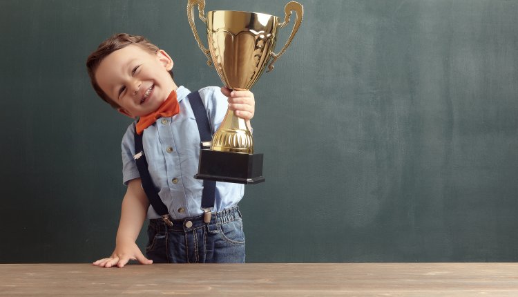 GPT Jongen met een grote glimlach houdt een gouden trofee vast terwijl hij naast een houten tafel staat voor een schoolbord. Hij draagt een lichtblauw overhemd met een oranje vlinderdas en bretels, en een paar donkerblauwe jeans. Zijn vreugde en trots zijn duidelijk zichtbaar.