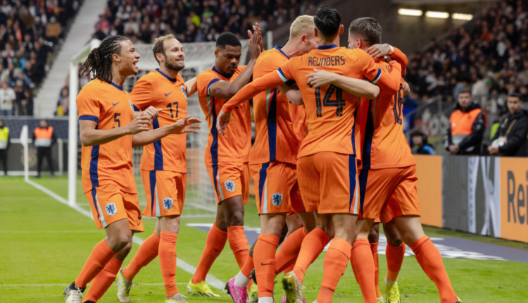 Nederlandse voetballers vieren een doelpunt tijdens een wedstrijd. Ze dragen oranje shirts met blauwe accenten. Een speler, te herkennen aan rugnummer 14, omhelst zijn teamgenoten terwijl anderen juichen. Op de achtergrond zijn toeschouwers en stadionpersoneel te zien.