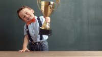 GPT Jongen met een grote glimlach houdt een gouden trofee vast terwijl hij naast een houten tafel staat voor een schoolbord. Hij draagt een lichtblauw overhemd met een oranje vlinderdas en bretels, en een paar donkerblauwe jeans. Zijn vreugde en trots zijn duidelijk zichtbaar.